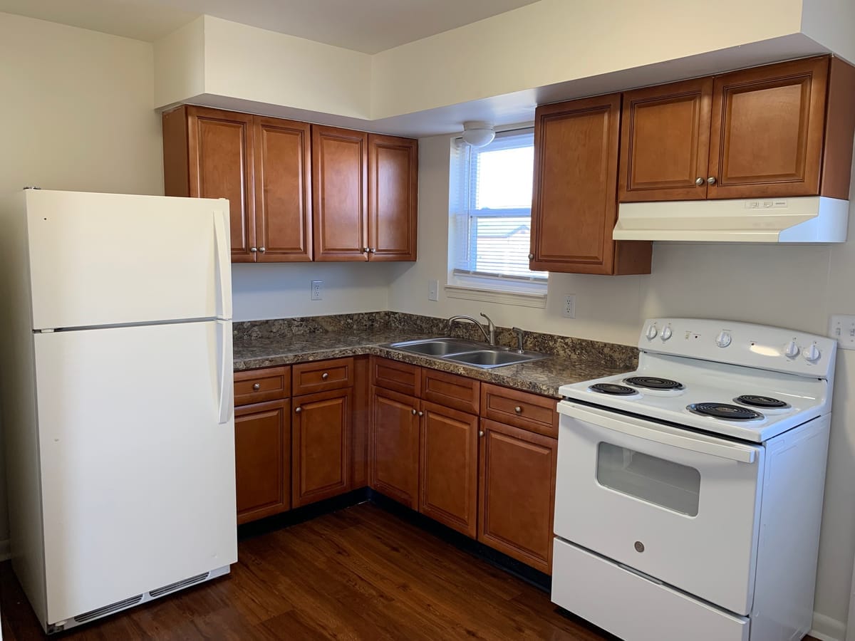 A Taneytown Village kitchen with wood cabinets, a white refrigerator and electric range, and a double sink below a window