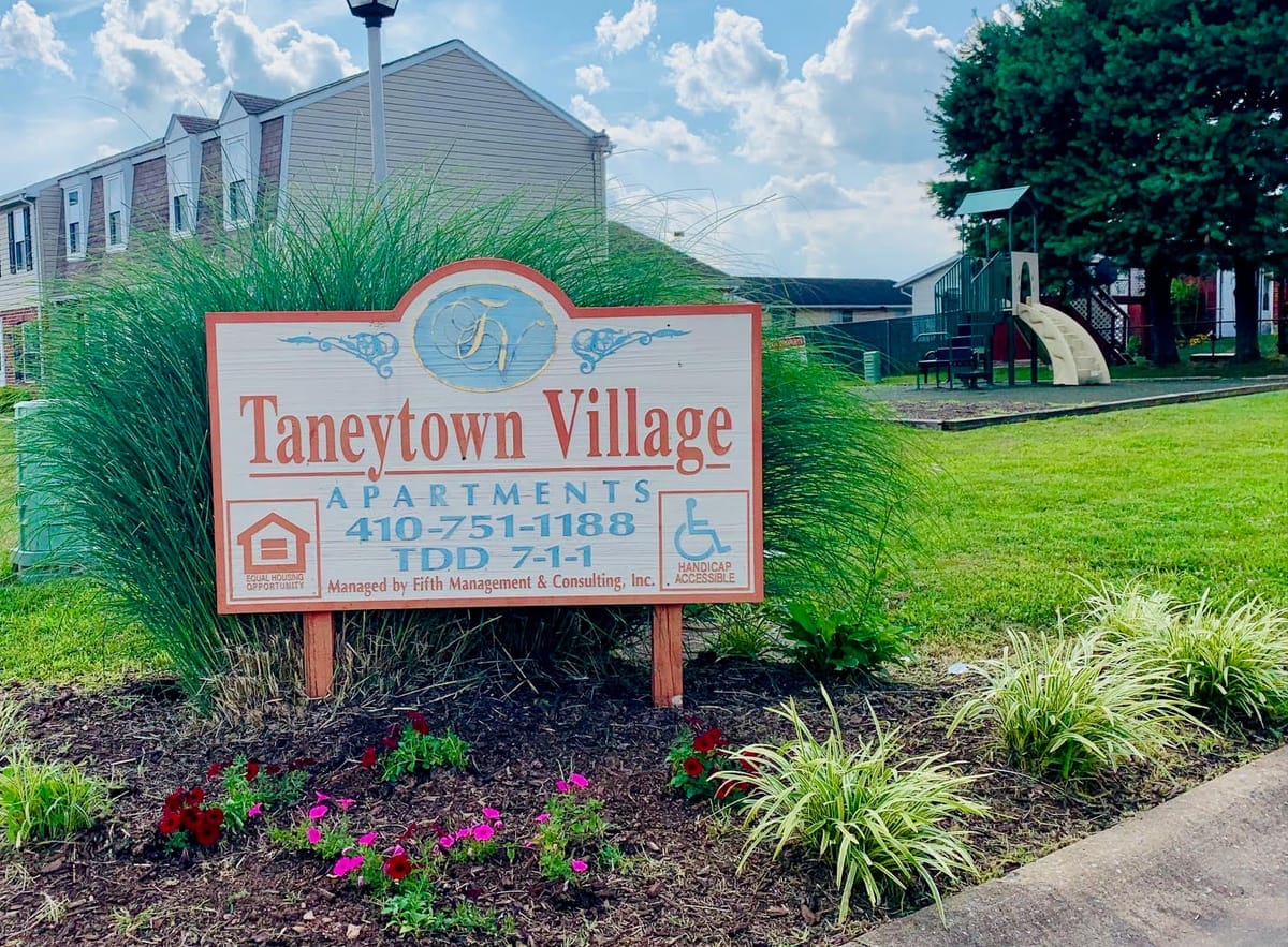 The Taneytown Village Apartments monument sign in a flower bed, with a townhouse and the on-site playground behind on a sunny day