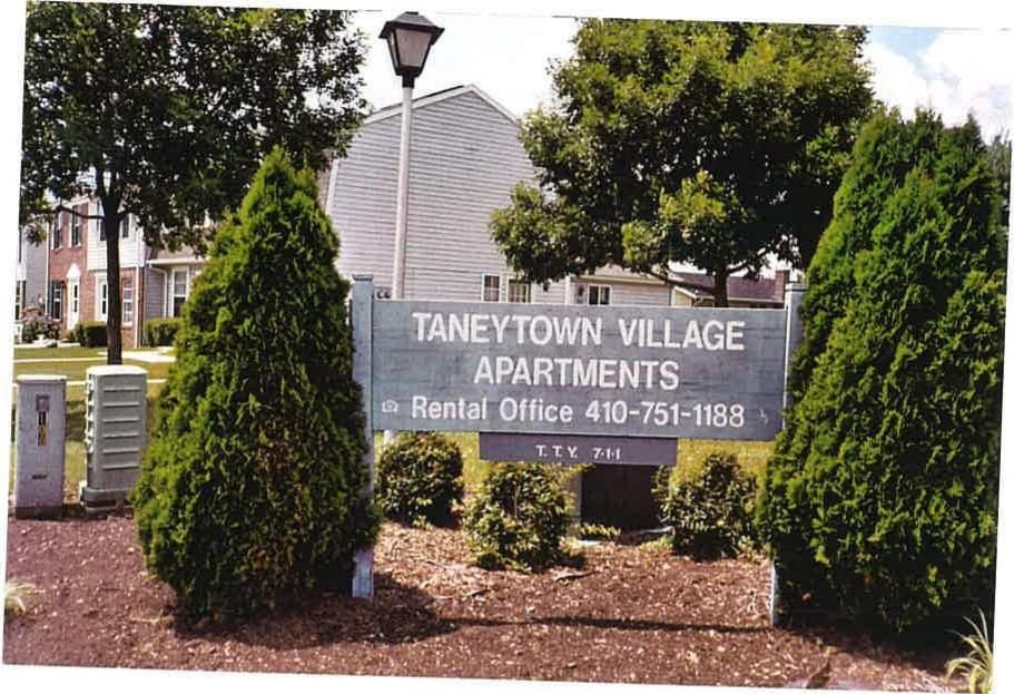 The Taneytown Village Apartments entrance sign among evergreen shrubs, with a townhouse behind